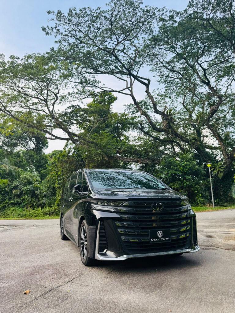 A sleek black Toyota Vellfire luxury van parked in a lush, natural setting with large trees, perfect for a premium chauffeur service