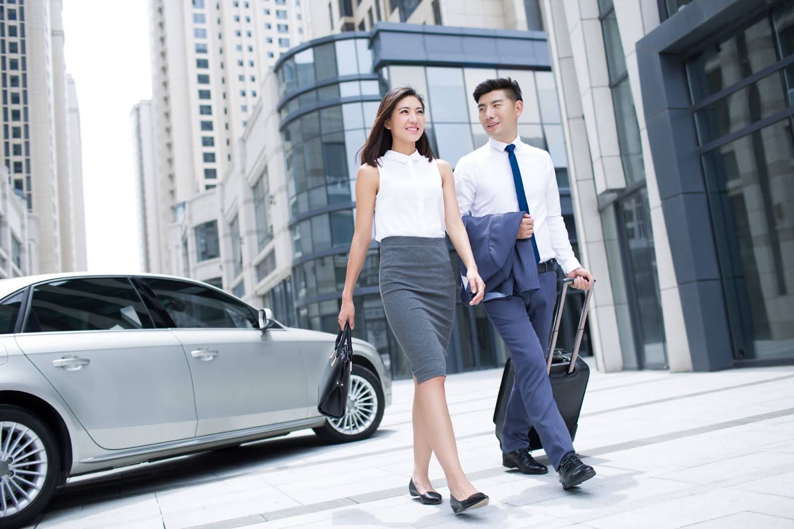 A professionally dressed man and woman walk side-by-side on a city sidewalk, surrounded by modern high-rise buildings.