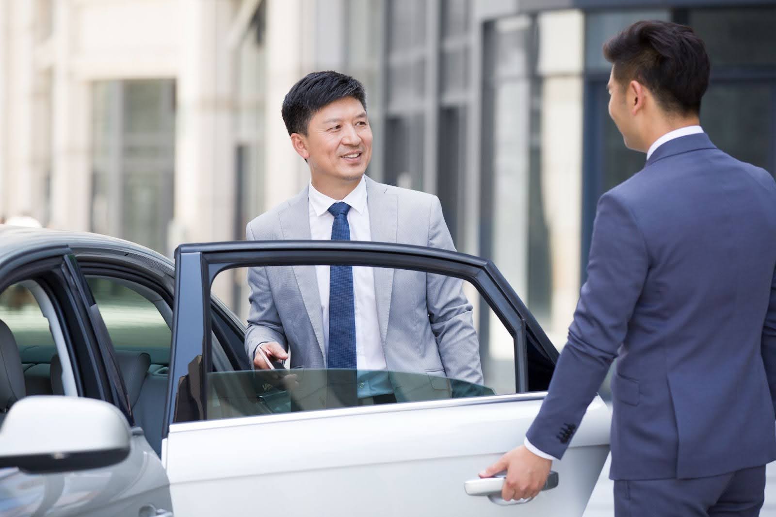 A chauffeur in a dark suit holds the door of a silver car open for a smiling businessman in a gray suit and tie who is getting out of the vehicle.