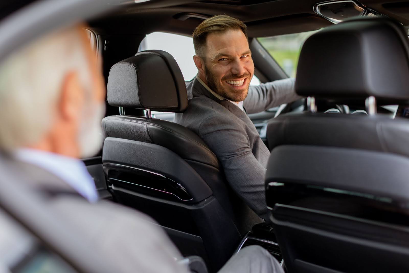 A man in a gray suit in the driver's seat of a car turns to smile warmly at his passenger, an older man with white hair who is seen from behind and out of focus.