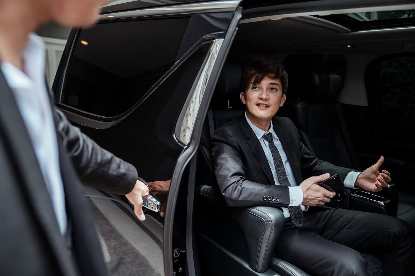 A smiling businessman in a suit holds a smartphone while sitting in the spacious back seat of a luxury MPV as a chauffeur opens the sliding door.