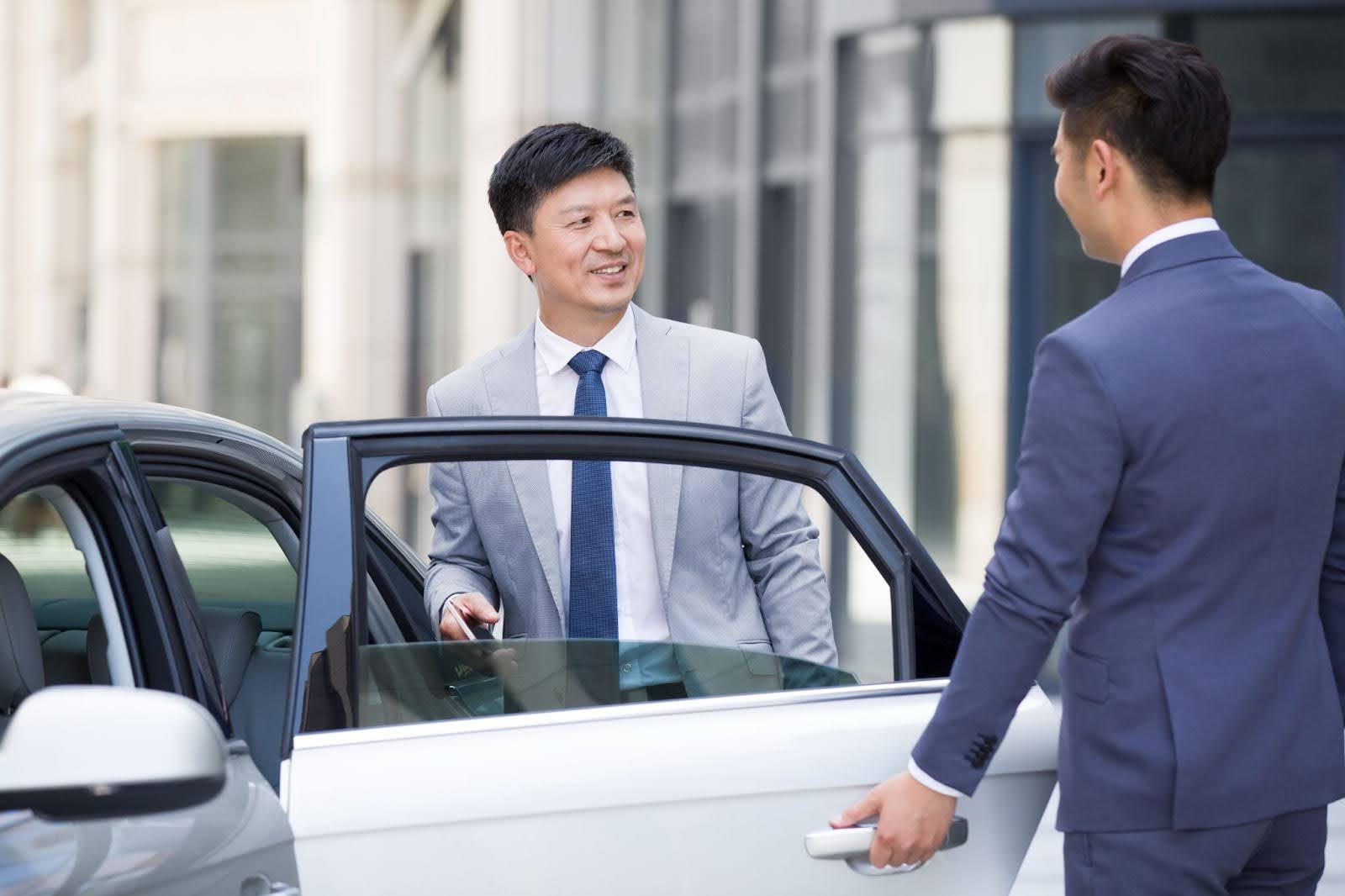 A professional chauffeur in a suit opens the door for a businessman exiting a luxury vehicle, illustrating the high-quality experience of a corporate limousine service
