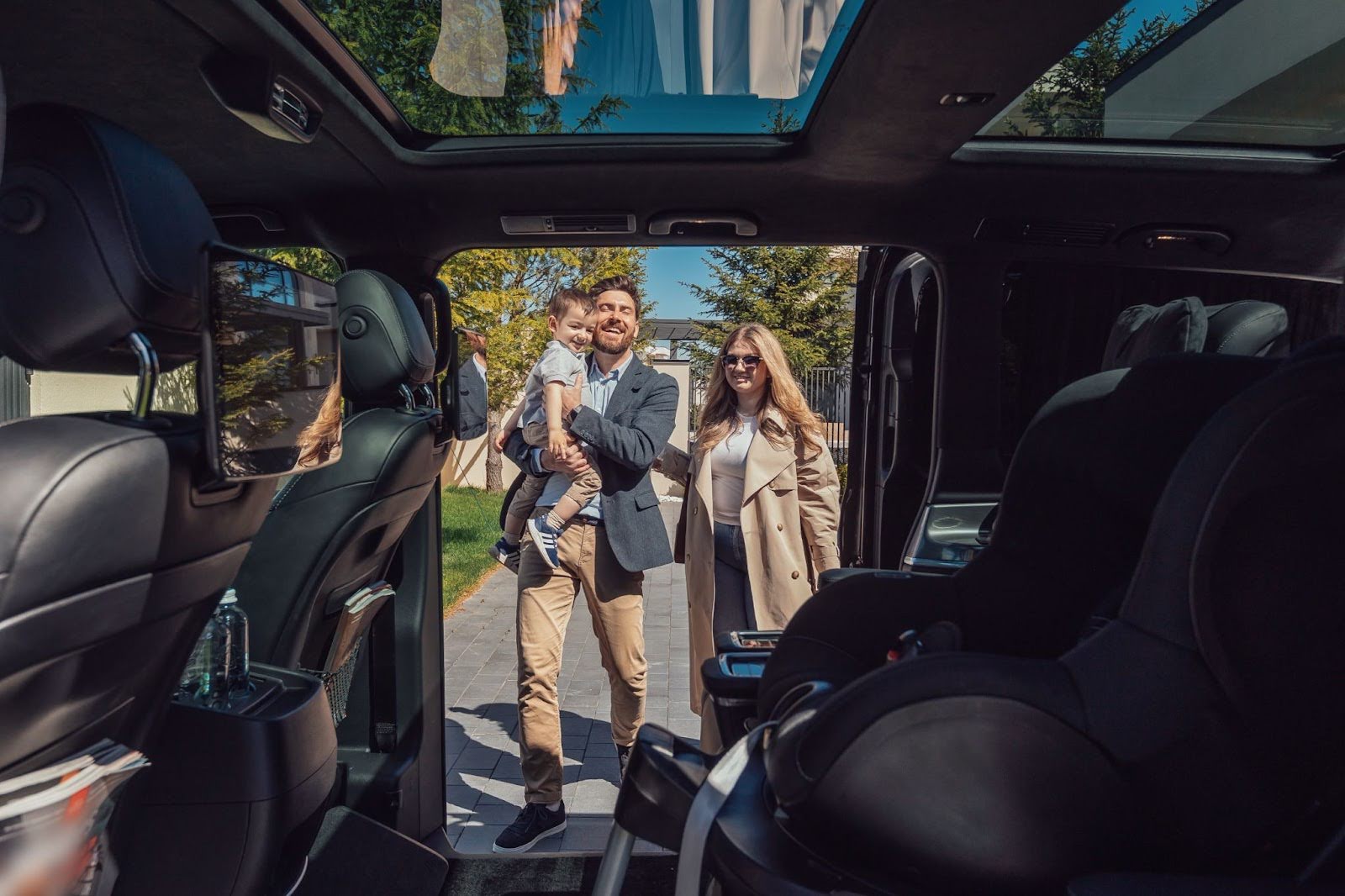 A smiling father carrying a toddler and a mother approach the open sliding door of a luxury van, viewed from the vehicle's interior which features a child safety seat.