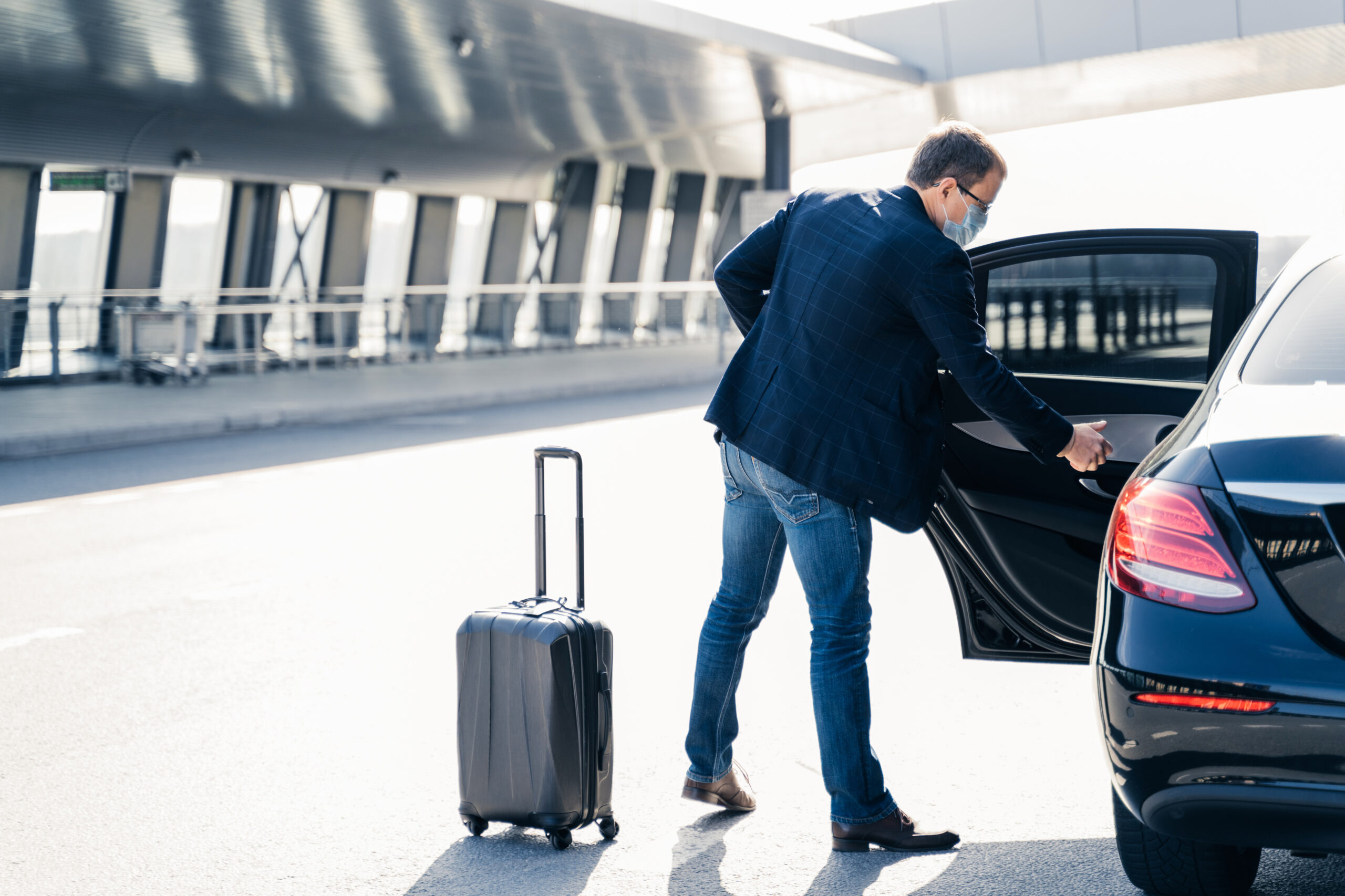 A traveler in a blue blazer and face mask opening the back door of a black car with a suitcase standing nearby at an airport terminal.