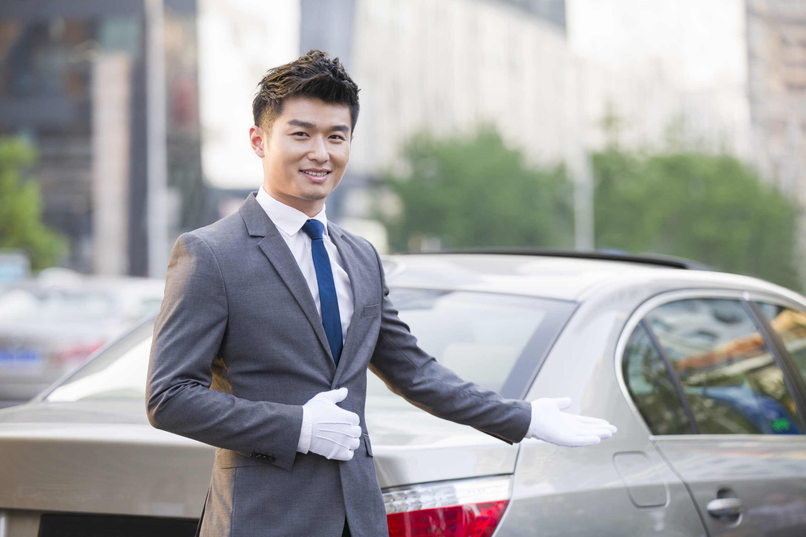 Smiling professional chauffeur wearing a grey suit and white gloves gesturing toward a luxury silver sedan