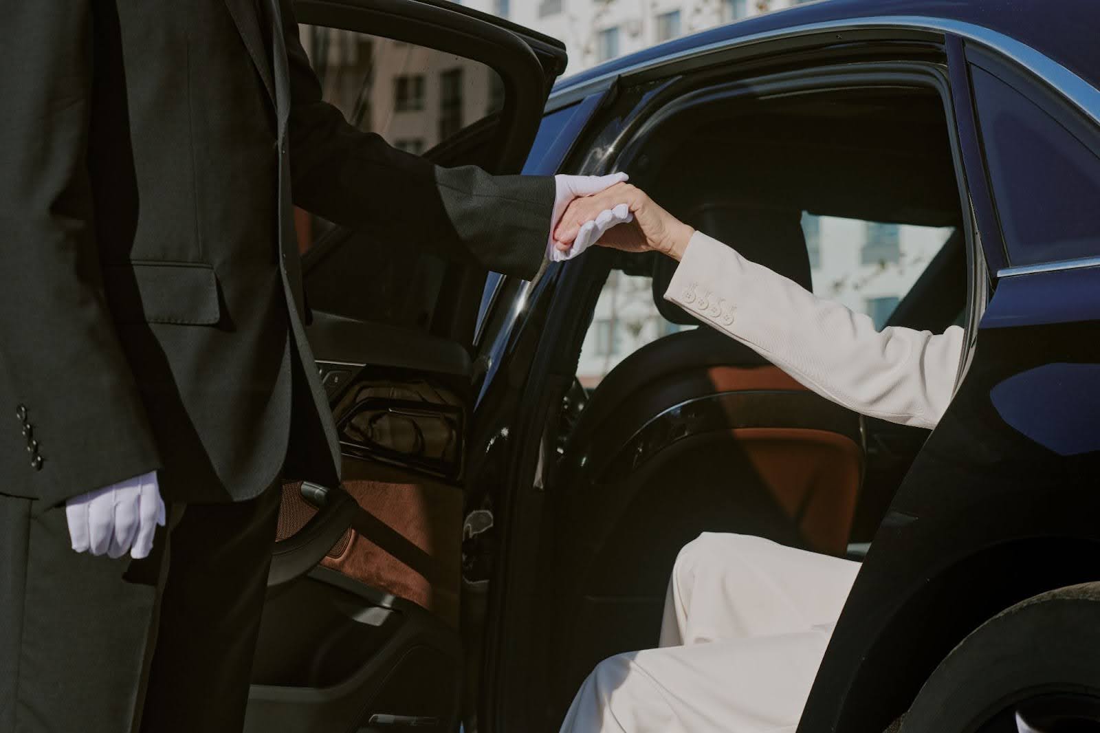 A professional chauffeur wearing white gloves assists a passenger in a white suit as they exit a luxury vehicle, showcasing premium corporate limousine service