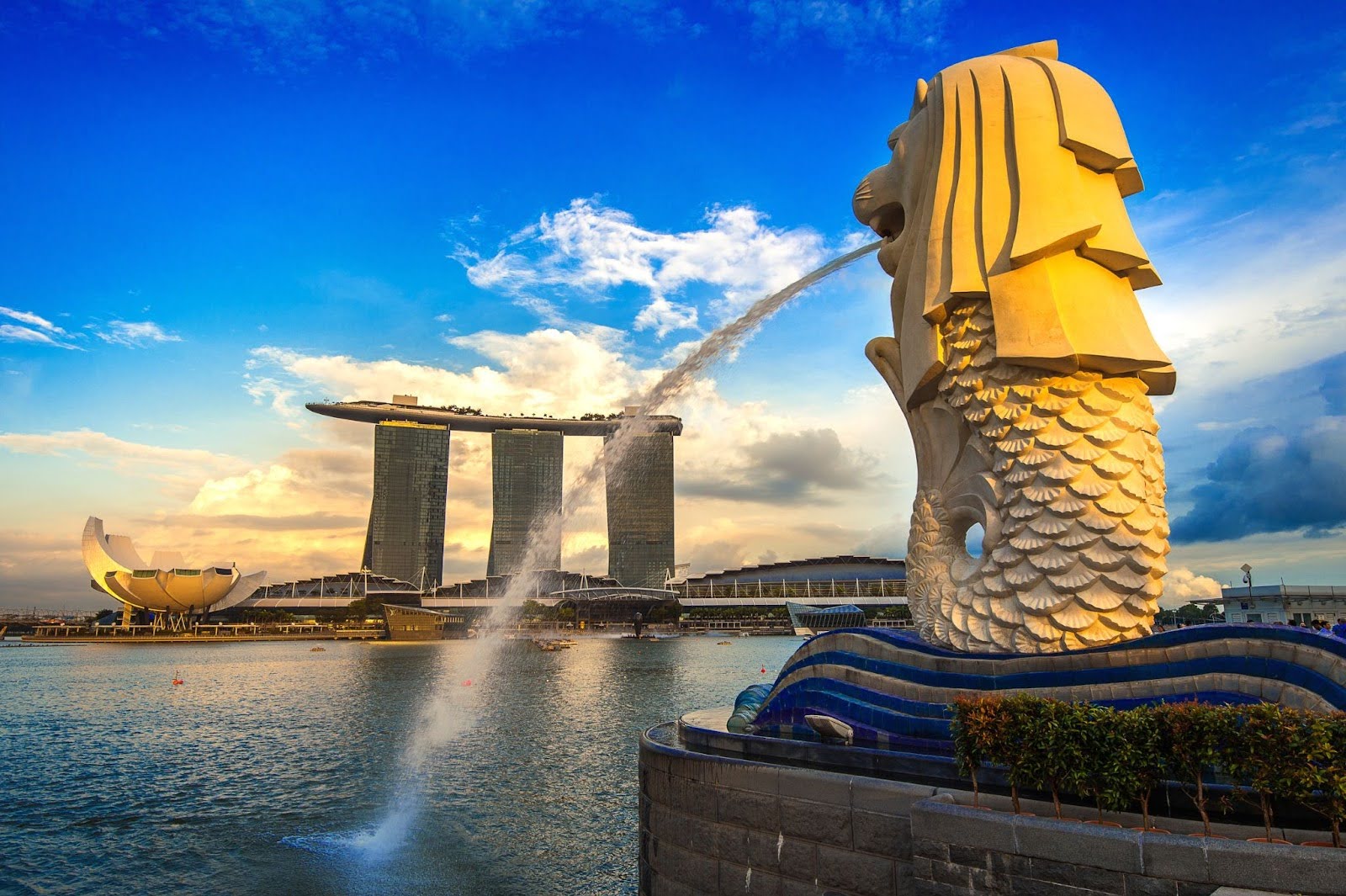 The iconic Merlion statue in Singapore spouts water into the bay at sunset, with the Marina Bay Sands hotel and the ArtScience Museum visible in the background against a vibrant sky.