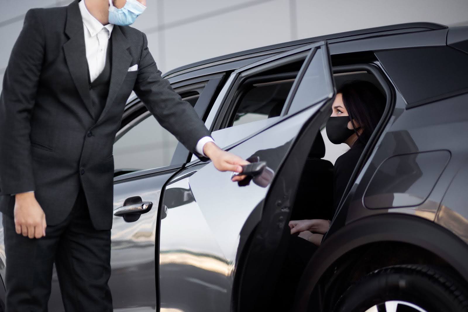 A professional chauffeur wearing a face mask opens the car door for a passenger utilizing a safe corporate limousine service.